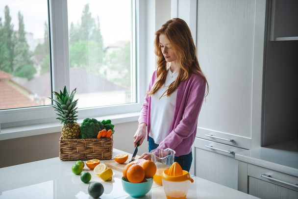 Widok z boku kobiety siedzącej w kuchni i krojącej wapno, pomarańcze, cytryny, grejpfruty na sok cytrusowy pełen witamin. Rude włosy i piegowata twarz - Zdjęcie, obraz