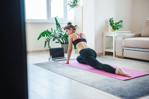 Zdolna kobieta w sportowej odzieży rozciągająca nogi, trenująca z laptopem, oglądająca samouczki online, ćwicząca pilates na macie do jogi. Trzymaj się. - Zdjęcie, obraz