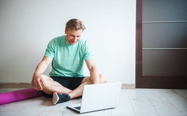 Mann wartet darauf, dass Online-Fitnesskurse beginnen, sitzt mit überkreuzten Beinen auf dem Boden und tippt etwas in seinen Laptop. Online-Schulungen. Online-Video - Foto, Bild