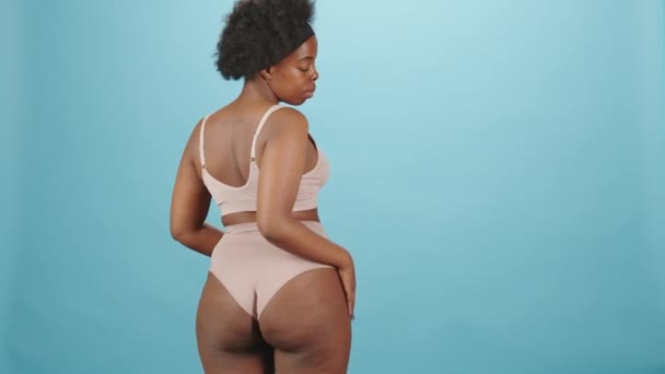 Medium achteraanzicht portret van mooie jonge bochtige Afro-Amerikaanse vrouw in roze ondergoed draaien om camera staan op geïsoleerde blauwe achtergrond - Video