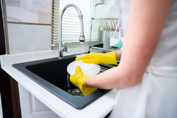 Sarı koruyucu eldivenli bir kadın mutfak lavabosunda bulaşıklarını yıkıyor. Ev işleri ve ev temizliği rutini. hizmetçi - Fotoğraf, Görsel
