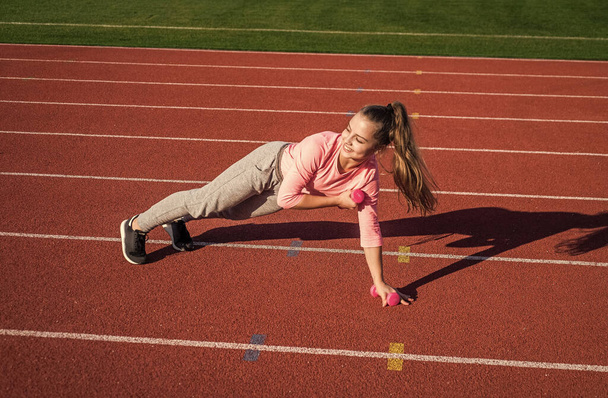 Power and balance. healthy childhood. workout on fresh air outdoor. confident athlete with barbell. teen girl warming up on stadium. kid in sportswear use dumbbell. child do exercise on racing track - Photo, Image