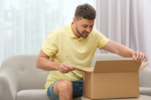 Happy man opening parcel on sofa at home. Internet shopping - Foto, imagen