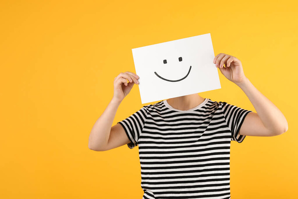 Woman hiding behind sheet of paper with happy face on yellow background, space for text - Foto, Imagem