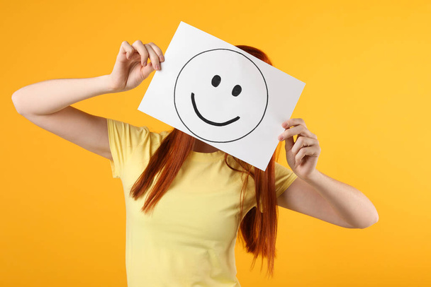 Woman hiding behind sheet of paper with happy face on yellow background - Foto, immagini