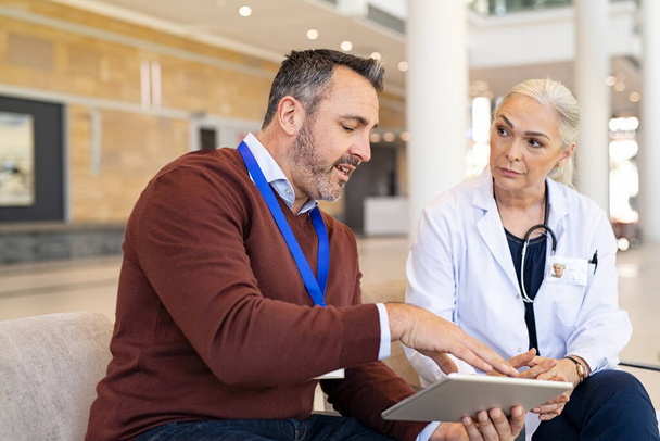 Pharmaceutical sales representative and mature woman physician having a discussion in hospital hallway. General practitioner discussing patient case status with his medical staff using digital tablet. Pharmaceutical representative showing medical rep - Foto, imagen