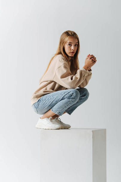 Cute-looking teenage girl crouching on a white cube, looking at the camera. In a studio over white background. - Fotoğraf, Görsel