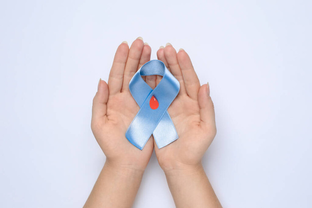 Woman holding light blue ribbon with drop of blood on white background, top view. Diabetes awareness - Фото, изображение