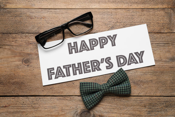 Card with phrase HAPPY FATHER'S DAY, eyeglasses and bow tie on wooden background, flat lay - Zdjęcie, obraz