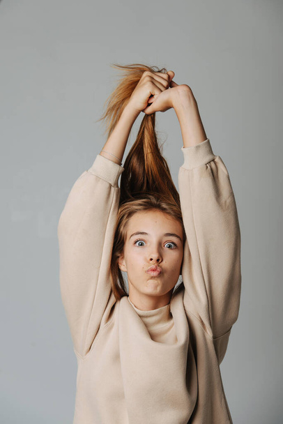 Funny grimacing teenage girl pulling her hair up, looking at the camera, making kissing lips. Against grey background. In a warm beige longsleeve. - Photo, image
