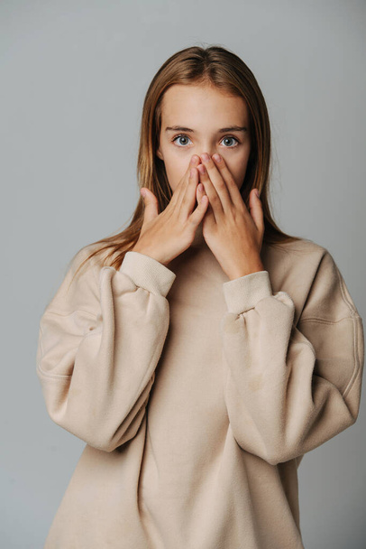 Surprised teenage girl covering her face with hands, looking at the camera. Against grey background. In a warm beige longsleeve. - Foto, afbeelding