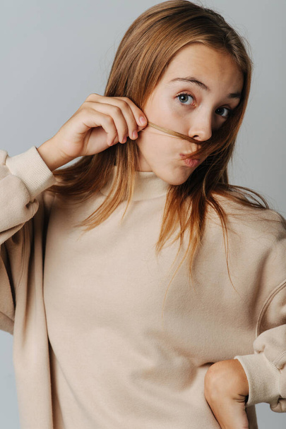 Cute pretty teenage girl playing with her har, making mustache, kissing lips grimace. Against grey background. In a warm beige longsleeve. - Photo, image