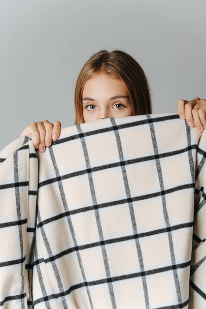 Timid teenage girl holding a checkered jacket over her face, peeking over it it. Over light grey background. - Foto, imagen