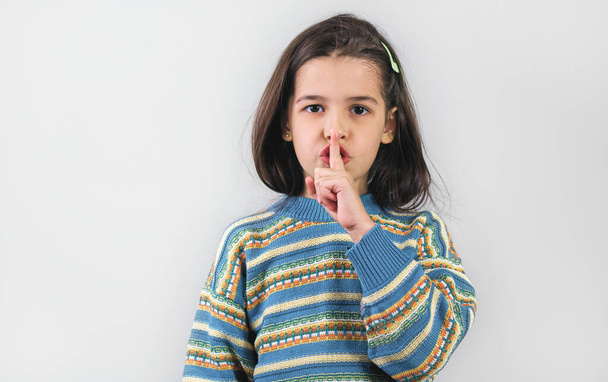 Studio portrait of little girl wearing colorful sweater showng quite gesture isolated over white background. Cute child making silence gesture. Kid holding finger on mouth shows shh sign. - Photo, image