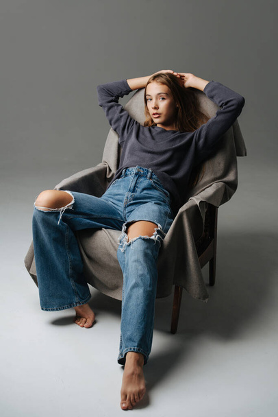 Chill teenage girl sitting on an armchair, holding hands over her head. Portrait, looking at the camera. Wearing mom jeans with holes on her knees and grey long-sleeve. - Foto, afbeelding