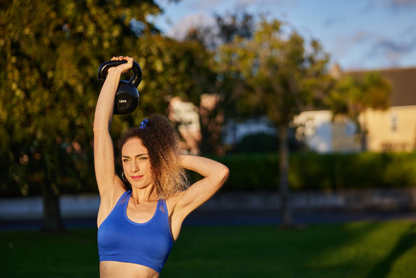 Caucasian blonde woman lifting a weight and exercising in a park - Foto, afbeelding