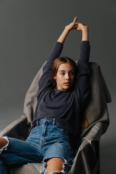 Bored, stretching on an armchair teenage girl, hands over her head. Portrait. Wearing mom jeans with holes and grey long-sleeve. - Photo, image