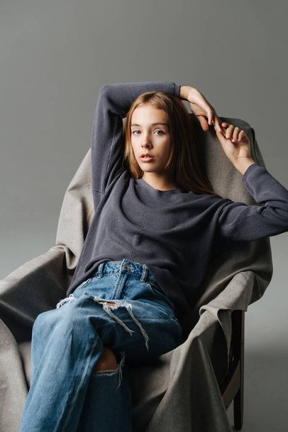 Sensual teenage girl sitting on an armchair, holding hands over her head. Portrait. Wearing mom jeans with holes and grey long-sleeve. - Фото, изображение