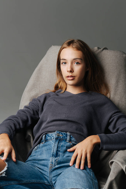Laid back teenage girl sitting on an armchair. Portrait, looking at the camera. Wearing mom jeans with holes and grey long-sleeve. - Fotoğraf, Görsel