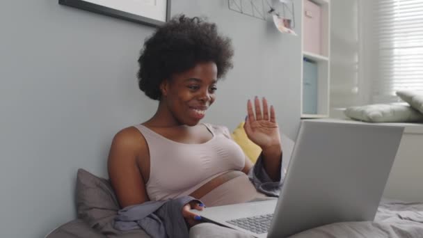 Medium slowmo shot of smiling young African-American woman having video chat on laptop while relaxing in bed at cozy apartment - Imágenes, Vídeo