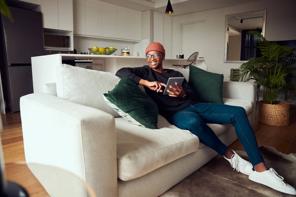 Adult black male smiling, looking out window while using at tablet. Modern apartment on the sofa - Foto, afbeelding