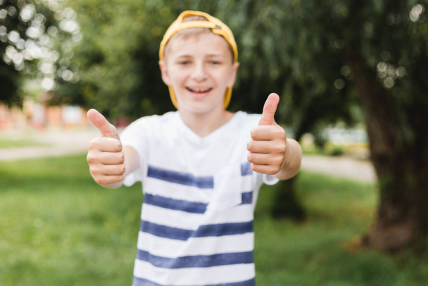 Muchacho sonrisa adolescente mostrando gesto pulgar hacia arriba en el fondo de la naturaleza, concepto de gesto. Niño vistiendo en camiseta de rayas y sombrero amarillo - Foto, Imagen