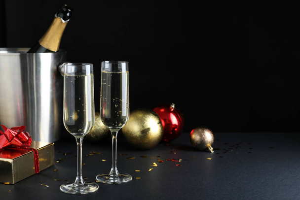 Happy New Year! Glasses with sparkling wine, bottle in bucket and festive decor on table against black background, space for text - Foto, Imagen