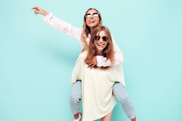 Two young beautiful smiling brunette hipster female in trendy summer clothes. Sexy carefree women posing near blue wall. Positive models having fun in sunglasses. Gives piggyback riding  - Photo, image