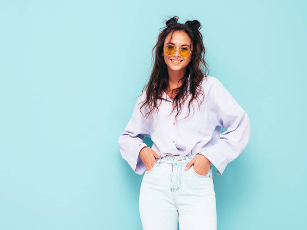 Young beautiful smiling female in trendy summer clothes. Sexy carefree woman posing near blue wall in studio. Positive brunette model having fun and going crazy. Cheerful and happy in sunglasses - Photo, Image