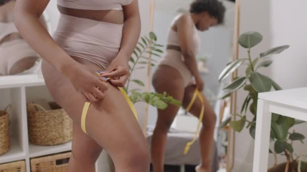 Keskiosa hidas laukaus nuori plus koko Afrikkalainen-amerikkalainen nainen alusvaatteet mittaavat hänen reidet keltainen mittanauha seisoo peilin edessä makuuhuoneessa - Materiaali, video