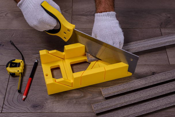A man cuts the skirting board using a saw and miter box on laminate floor. - Zdjęcie, obraz
