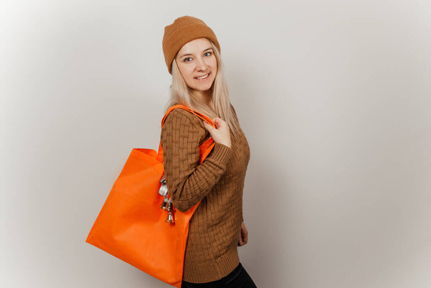 Frau in braunem Pullover und Hut mit orangefarbener Einkaufstasche vor grauem Hintergrund - Foto, Bild