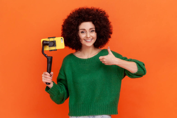 Smiling woman with Afro hairstyle wearing green casual style sweater using steadicam and smart phone, making video for her vlog, showing thumb up. Indoor studio shot isolated on orange background. - Foto, immagini