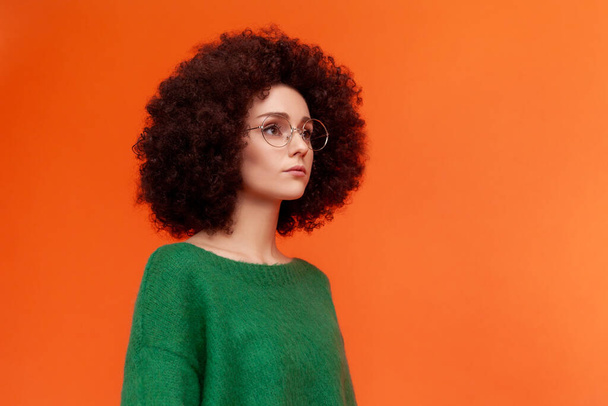 Profile portrait of good looking woman with Afro hairstyle wearing green casual style sweater and eyeglasses, looking away, serious expression. Indoor studio shot isolated on orange background. - Fotografie, Obrázek