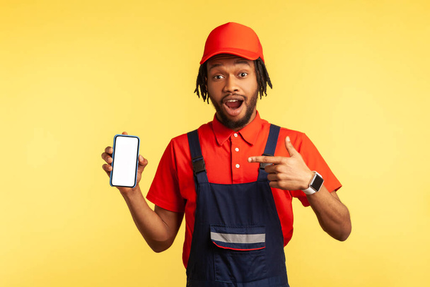 Shocked workman in overalls pointing at mobile phone with blank screen, surprised by online delivery order app, house repair maintenance services. Indoor studio shot isolated on yellow background. - Foto, immagini