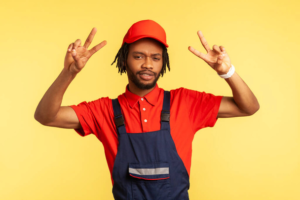 Positive handyman with beard in overalls looking at camera, showing v sign, working in service industry, courier delivery, housekeeping maintenance. Indoor studio shot isolated on yellow background. - Fotografie, Obrázek