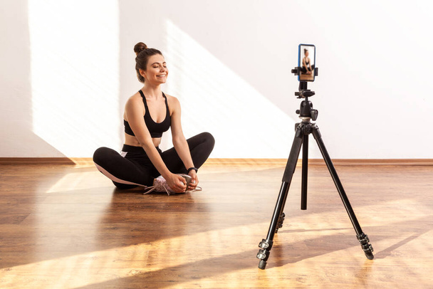 Female coach filming video of relaxing after workout, sitting on lotus pose with closed eyes, wearing black sports top and tights. Full length studio shot illuminated by sunlight from window. - Foto, imagen