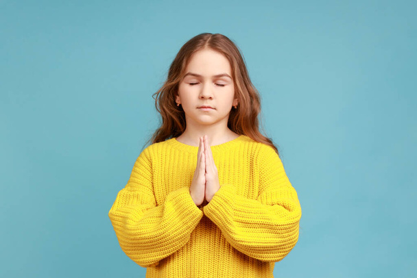 Portrait of little girl holding fingers in mudra gesture, feeling calm positive and relaxed, yoga practice, wearing yellow casual style sweater. Indoor studio shot isolated on blue background. - Fotó, kép