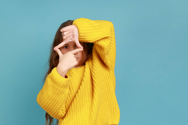 Portrait of little girl looking through photo frame hand gesture, focusing picture, observing world, wearing yellow casual style sweater. Indoor studio shot isolated on blue background. - Foto, afbeelding