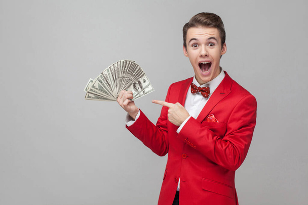 Wow, look at my money! Portrait of surprised man with stylish hairdo in red jacket and bow tie pointing at bunch of dollars, expressing extreme joy and amazement. indoor studio shot, gray background - Foto, imagen
