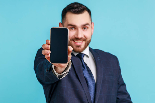 Online service, technology. Satisfied happy man wearing official style suit standing holding out cellphone and smiling broadly at camera. Indoor studio shot isolated on blue background. - Photo, image
