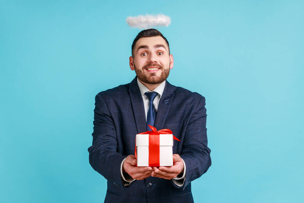 Bearded man with kind smile wearing official style suit and halo overhead holding gift box, charity organization, religion, looking at camera. Indoor studio shot isolated on blue background. - Zdjęcie, obraz