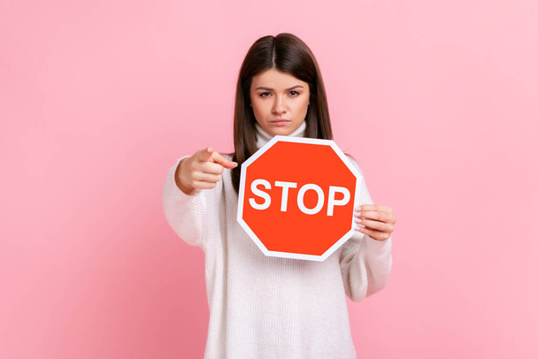 Strict bossy woman pointing to camera and holding Stop road traffic sign as symbol of prohibition, wearing white casual style sweater. Indoor studio shot isolated on pink background. - Fotó, kép