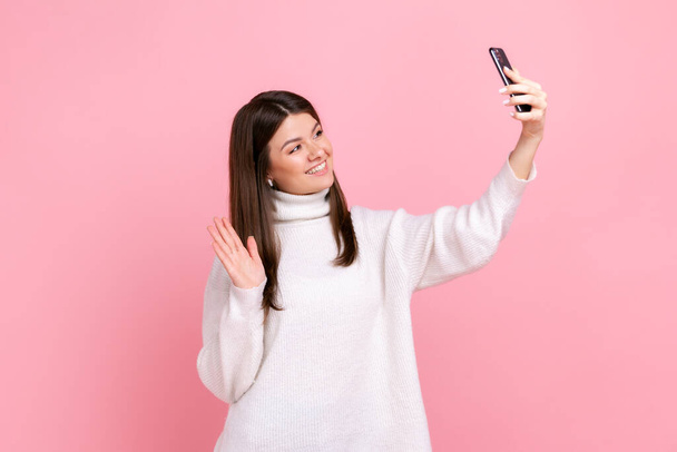 Woman taking selfie and waving hand, gesturing hello for followers, communicating on video call, wearing white casual style sweater. Indoor studio shot isolated on pink background. - Photo, Image