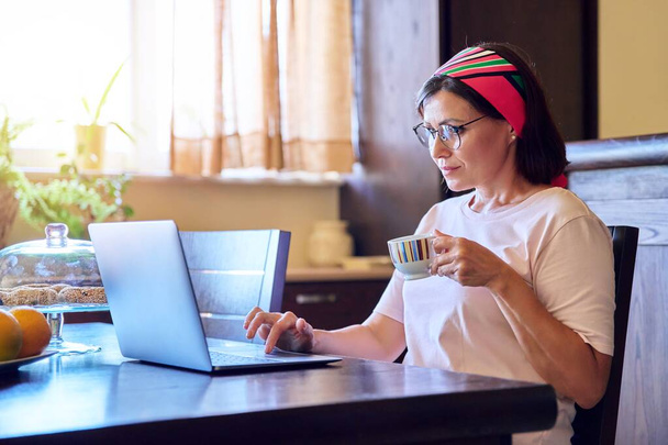 Mature woman relaxing at home at the table in the living room with a laptop and a cup of coffee - Фото, зображення