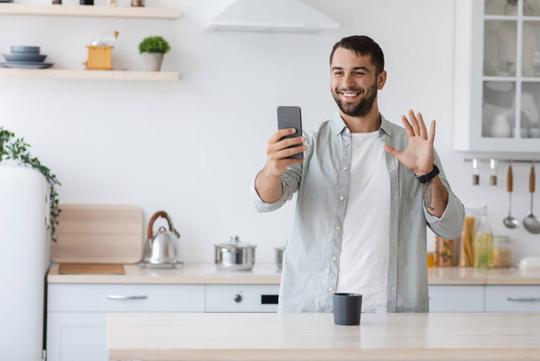 Cheerful handsome adult european male waving hand to smartphone webcam, making video calls from phone - Fotoğraf, Görsel