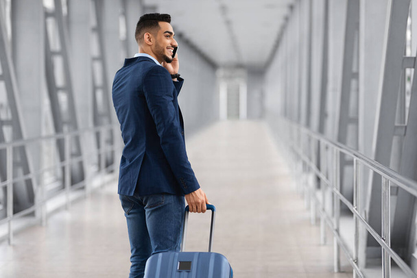 Después de la llegada. Un hombre de negocios árabe sonriente hablando por celular mientras pasaba por la puerta del aeropuerto - Foto, Imagen