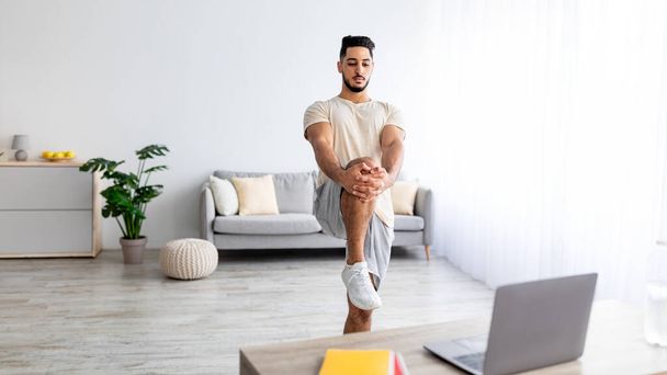 Hombre árabe joven estirando su pierna delante de la computadora portátil durante el entrenamiento doméstico, siguiendo video tutorial en línea en la computadora portátil - Foto, Imagen