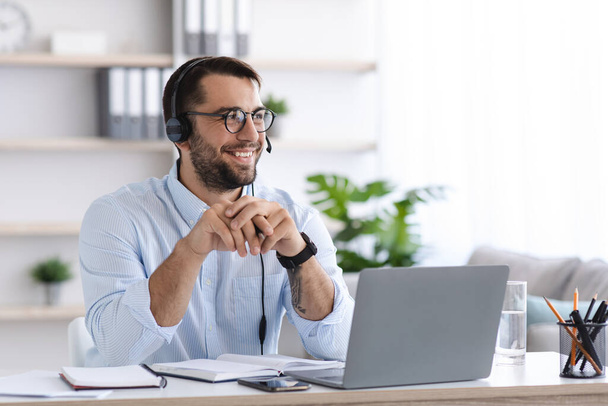Взрослый европейский мужчина с бородой в очках, наушниках работает на ноутбуке, разговаривает с клиентами в офисе или в интерьере комнаты - Фото, изображение