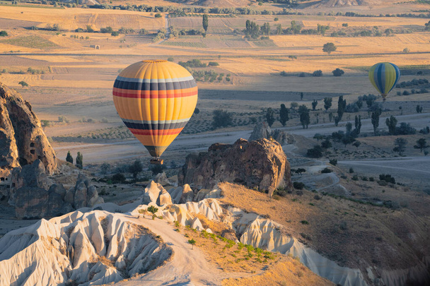 landscape with lonely hot air balloon with rainbow colors pattern rising over the Cappadocian valley panorama in the morning light with chimney rock formations in the foreground - Foto, Imagen